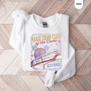May include: White sweatshirt with a colorful graphic featuring a monorail, fireworks, and the text "PLEASE STAND CLEAR of the Doors." The design includes a sphere and the text "Por Favor, Mantingase Alejado De Las Puertas."