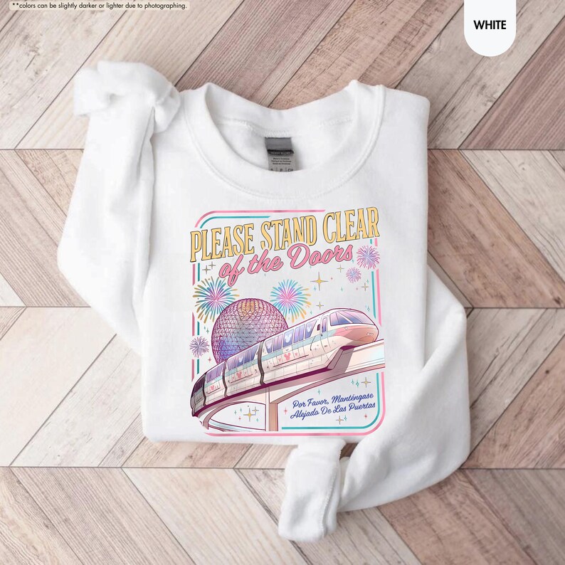 May include: White sweatshirt with a colorful graphic featuring a monorail, fireworks, and the text "PLEASE STAND CLEAR of the Doors." The design includes a sphere and the text "Por Favor, Mantingase Alejado De Las Puertas."