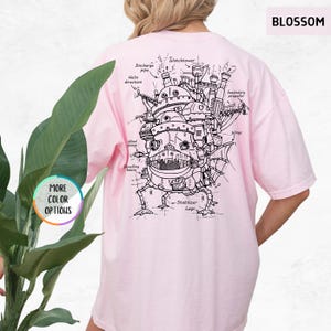 Op de afbeelding: Lichtroze t-shirt met een gedetailleerde zwart-wit illustratie van een bewegend kasteel, met gelabelde componenten zoals "Watchtower" en "Stabilizer Legs". Het woord "BLOSSOM" staat rechtsboven gedrukt. Het shirt heeft ook de tekst "MORE COLOR OPTIONS".