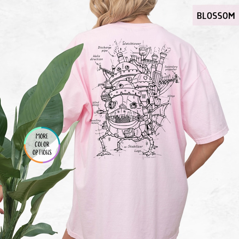 Op de afbeelding: Lichtroze t-shirt met een gedetailleerde zwart-wit illustratie van een bewegend kasteel, met gelabelde componenten zoals "Watchtower" en "Stabilizer Legs". Het woord "BLOSSOM" staat rechtsboven gedrukt. Het shirt heeft ook de tekst "MORE COLOR OPTIONS".