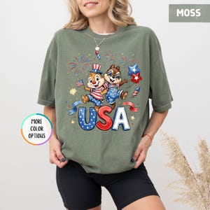 May include: Moss green t-shirt featuring a graphic of two cartoon chipmunks celebrating with fireworks and the word "USA" in red and blue. The chipmunks are dressed in patriotic outfits, holding a flag and star-shaped balloons.