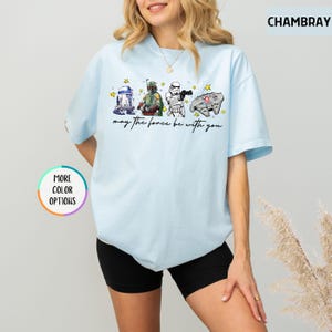 Puede incluir: Camiseta azul claro con personajes de Star Wars: R2-D2, Boba Fett, un Stormtrooper y el Halc&oacute;n Milenario. La frase "may the force be with you" est&aacute; impresa debajo. La camiseta tiene mangas cortas y cuello redondo.