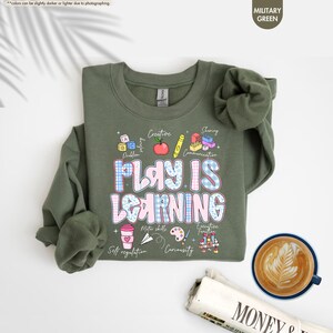 May include: A military green sweatshirt with the text "PLAY IS LEARNING" in a blue and pink plaid design. The sweatshirt features illustrations of an apple, pencil, and other educational icons. A cup of coffee and a newspaper are in the background.