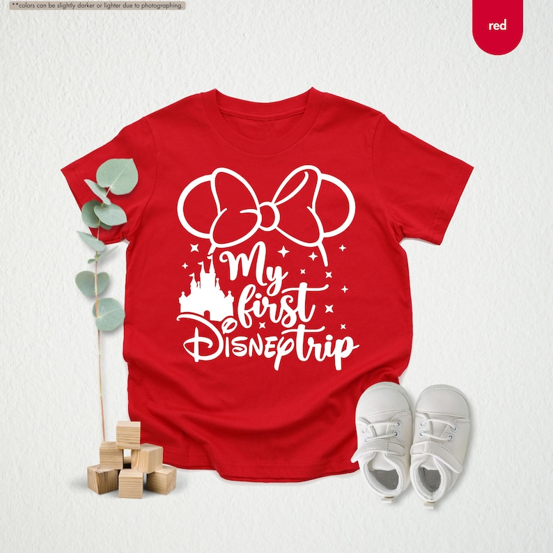 Pu&ograve; includere: Maglietta rossa con una grafica bianca di un castello, orecchie da topo con un fiocco e il testo "My first DISNEY trip".