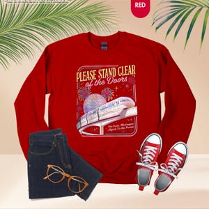 May include: A red sweatshirt with the text "PLEASE STAND CLEAR of the Doors" and a graphic of a monorail and fireworks. The image also includes a pair of blue jeans, gold-rimmed glasses, and red sneakers.