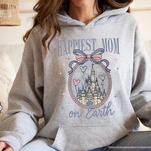 May include: Sport grey hoodie with a front graphic that reads "Happiest Mom on Earth." The graphic features a castle design with a pink and blue bow and Mickey Mouse balloons. The hoodie has a drawstring hood and a front pocket.
