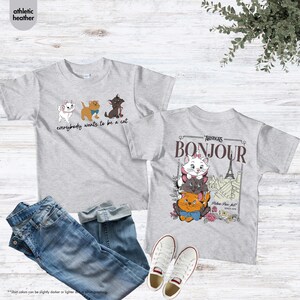 May include: Two light gray t-shirts with cat graphics. One shirt says "everybody wants to be a cat" with three cartoon cats. The other shirt says "Bonjour" with a stack of cats and the Eiffel Tower. Blue jeans and white sneakers are also in the image.