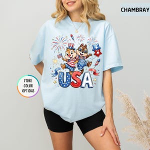May include: Light blue t-shirt featuring a cartoon illustration of two chipmunks celebrating with fireworks and the word "USA". The chipmunks are wearing patriotic outfits. The shirt has short sleeves and a round neckline.
