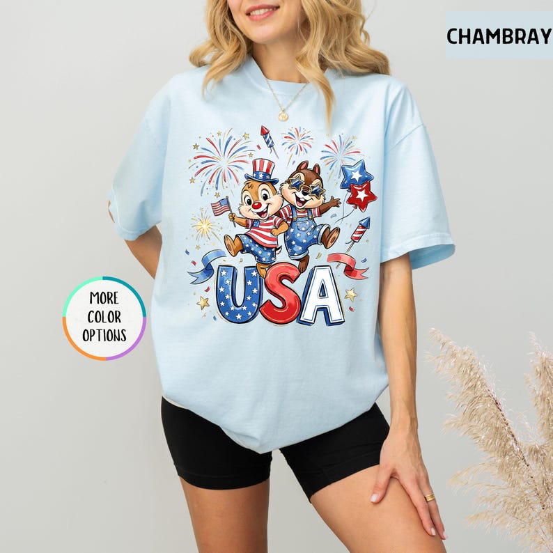 May include: Light blue t-shirt featuring a cartoon illustration of two chipmunks celebrating with fireworks and the word "USA". The chipmunks are wearing patriotic outfits. The shirt has short sleeves and a round neckline.
