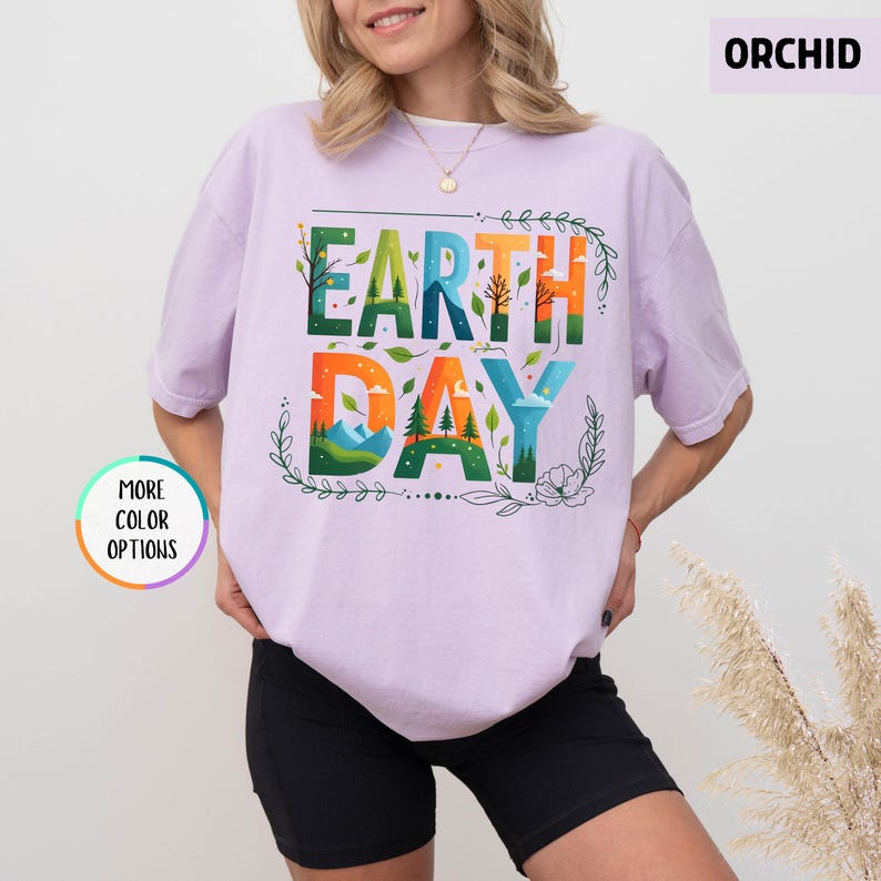 May include: A light purple t-shirt with the text "EARTH DAY" in colorful letters surrounded by green leaves and flowers. The text is in a stylized font with a layered effect.