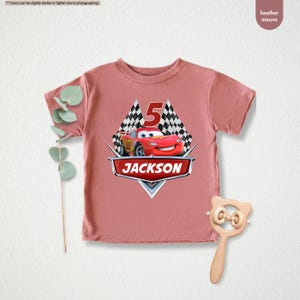 Op de afbeelding: Een heidepaars T-shirt met een rode raceauto-afbeelding en de tekst "JACKSON" en "5" erop gedrukt.