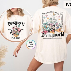 Puede incluir: Camiseta color marfil con dise&ntilde;o de Disney World. El gr&aacute;fico frontal incluye "Disneyworld" sobre personajes de Disney y "Florida Est. 1971." La parte trasera muestra un gr&aacute;fico m&aacute;s grande de Disney World con personajes y el texto "Disneyworld 1971."