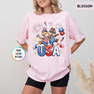 May include: Light pink t-shirt featuring a graphic of two cartoon chipmunks celebrating with fireworks and the word "USA" in red, white, and blue. The shirt has short sleeves and a crew neck.