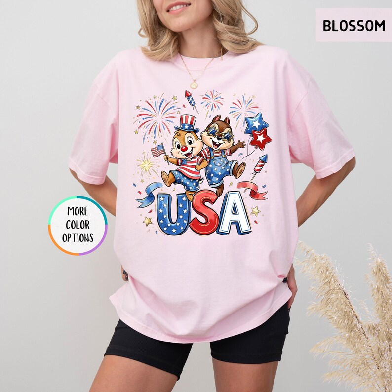 May include: Light pink t-shirt featuring a graphic of two cartoon chipmunks celebrating with fireworks and the word "USA" in red, white, and blue. The shirt has short sleeves and a crew neck.