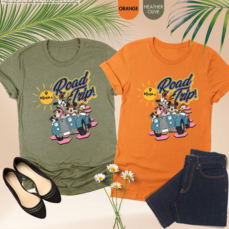 Op de afbeelding: Twee T-shirts, &eacute;&eacute;n oranje en &eacute;&eacute;n olijfgroen, met een afbeelding van Mickey Mouse, Minnie Mouse, Goofy en Donald Duck in een blauwe auto met de tekst "Road Trip! Miami". Een paar zwarte ballerina's en een blauwe jeans staan ook op de foto.