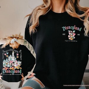 May include: Black sweatshirt with a white graphic print of Mickey Mouse characters in front of the Disneyland castle. The text "Disneyland Est 1955 California" is printed below the graphic.
