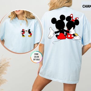 May include: Pale blue t-shirt with Mickey and Minnie Mouse graphics. The front shows the characters kissing, and the back has a larger image of them embracing. The word "CHAMBRAY" is printed on the top right of the t-shirt.
