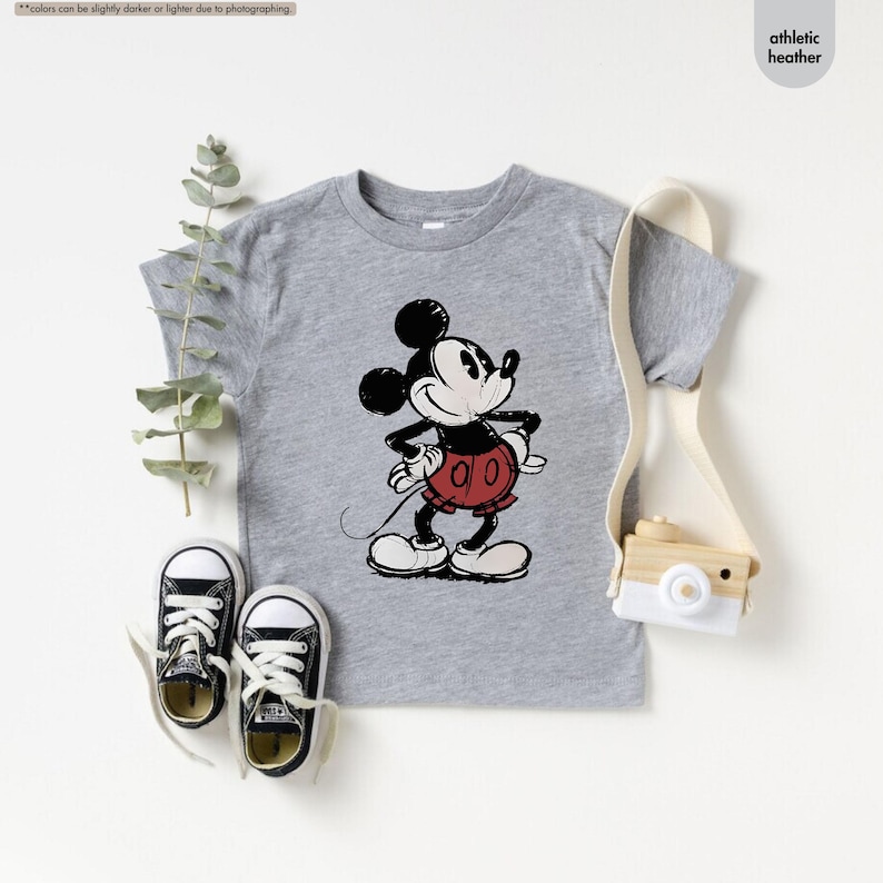 May include: A gray athletic heather t-shirt featuring a distressed graphic of Mickey Mouse. Mickey is depicted in black, white, and red, with his hands on his hips. The shirt is paired with black canvas sneakers.