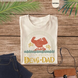 Puede incluir: Una camiseta beige con una ilustraci&oacute;n de estilo vintage de un personaje de dibujos animados con un cuerpo rojo y una corona amarilla. El texto "Just an ordinary Demi-Dad" est&aacute; impreso debajo de la ilustraci&oacute;n.