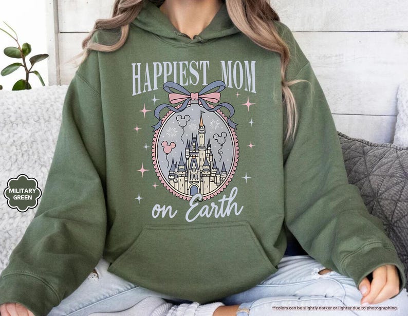 May include: Military green hoodie with a front graphic that reads "Happiest Mom on Earth." The graphic features a castle, balloons, and stars within a circular frame, with a bow at the top. The hoodie has a kangaroo pocket and a drawstring hood.