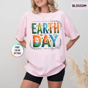 May include: A pink t-shirt with the words "Earth Day" in colorful letters. The letters are made up of images of nature, such as trees, mountains, and clouds.