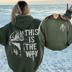 May include: A green hooded sweatshirt with a white graphic print of a Mandalorian helmet and the text "This is the Way".