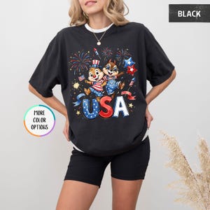 May include: Black t-shirt featuring a graphic of two cartoon chipmunks celebrating with fireworks and the word "USA". The chipmunks are wearing patriotic outfits. The shirt has a crew neck and short sleeves.
