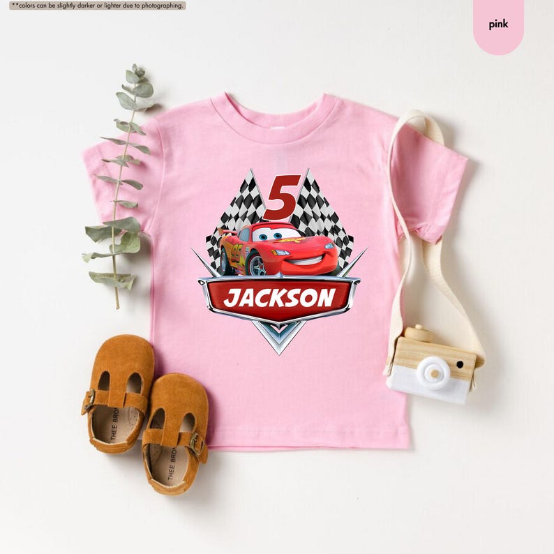 Op de afbeelding: Roze T-shirt met een rode Lightning McQueen auto-afbeelding, geblokte vlaggen en het nummer 5. De naam "JACKSON" is gedrukt in een zilveren banner onder de auto.