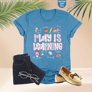 May include: A teal t-shirt with the words "PLAY IS LEARNING" in a colourful font. The shirt is styled with dark denim jeans, a pair of glasses, and cream-coloured loafers. The shirt also has small graphics and words like "Creative" and "Motor skills".