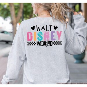 Puede incluir: Sudadera gris claro con la inscripci&oacute;n "WALT DISNEY WORLD" en letras coloridas en la espalda. El texto incluye un patr&oacute;n de cuadros blancos y negros y siluetas de Mickey Mouse.