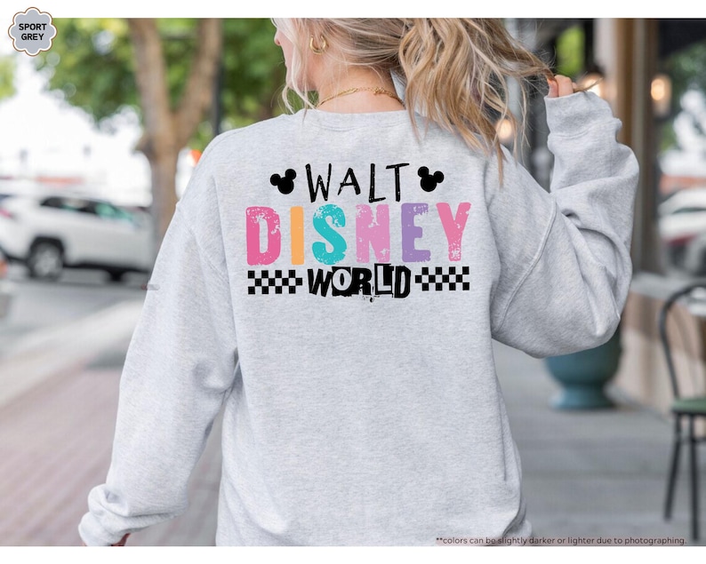 Puede incluir: Sudadera gris claro con la inscripci&oacute;n "WALT DISNEY WORLD" en letras coloridas en la espalda. El texto incluye un patr&oacute;n de cuadros blancos y negros y siluetas de Mickey Mouse.