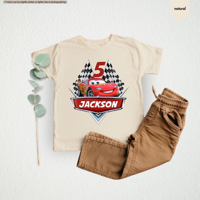 Op de afbeelding: Een beige T-shirt met een rode raceauto en geblokte vlaggen. Het nummer 5 staat op de vlaggen. De tekst "JACKSON" staat op het shirt.