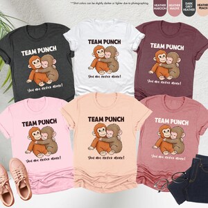 Puede incluir: Una colecci&oacute;n de camisetas en varios colores, incluyendo gris jaspeado, blanco y rosa. Cada camiseta presenta el texto "TEAM PUNCH" sobre una ilustraci&oacute;n de dos monos abraz&aacute;ndose, con la frase "You are never alone!" debajo.