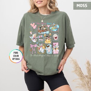 May include: Moss green t-shirt featuring a colorful graphic of Disney characters and icons. The design includes Mickey and Minnie Mouse, Dumbo, and other elements. Text on the shirt reads "Walt Disney World" and "The Most Magical Place on Earth."