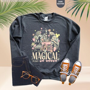 Pode incluir: Camisola cinza escuro com um gr&aacute;fico colorido que diz "The Most Magical Place". O design inclui um castelo, orelhas do Mickey Mouse e outros elementos do parque tem&aacute;tico. A imagem tamb&eacute;m mostra jeans escuros, t&ecirc;nis laranja e &oacute;culos.