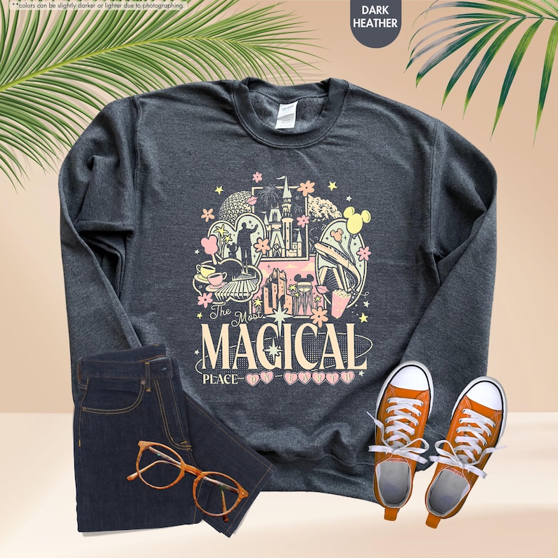 Pode incluir: Camisola cinza escuro com um gr&aacute;fico colorido que diz "The Most Magical Place". O design inclui um castelo, orelhas do Mickey Mouse e outros elementos do parque tem&aacute;tico. A imagem tamb&eacute;m mostra jeans escuros, t&ecirc;nis laranja e &oacute;culos.