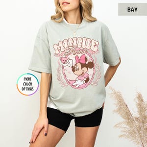 May include: A light green t-shirt with a pink and white graphic of Minnie Mouse holding a teacup. The graphic says "Minnie Mouse" in a retro font.
