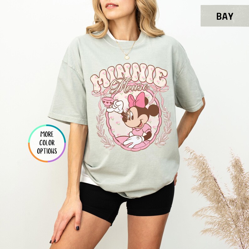 May include: A light green t-shirt with a pink and white graphic of Minnie Mouse holding a teacup. The graphic says "Minnie Mouse" in a retro font.