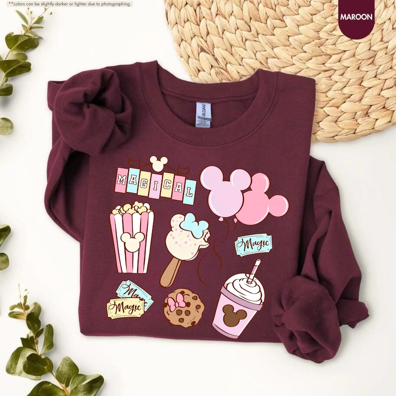 Puede incluir: Una sudadera color granate con un estampado gr&aacute;fico de art&iacute;culos con tem&aacute;tica de Mickey Mouse, que incluyen palomitas de ma&iacute;z, globos, helado, una galleta y una bebida. El texto "MAGICAL" y "Magic" tambi&eacute;n se incluyen en el dise&ntilde;o.