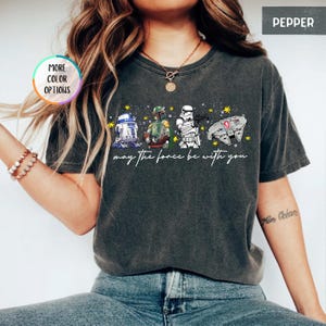 Puede incluir: Camiseta gris oscuro con los personajes de Star Wars R2-D2, Boba Fett, un Stormtrooper y el Halc&oacute;n Milenario. La frase "may the force be with you" est&aacute; escrita debajo. La camiseta tiene un aspecto vintage y desgastado.