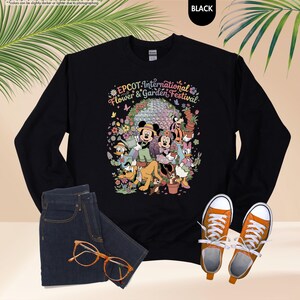 May include: Black sweatshirt featuring a colorful graphic of Disney characters and the text "EPCOT International Flower & Garden Festival." The design includes Mickey and Minnie Mouse, Donald Duck, and other characters. The sweatshirt is paired with blue jeans and orange sneakers.