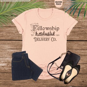 Puede incluir: Una camiseta de color melocot&oacute;n con un gr&aacute;fico marr&oacute;n de los personajes de la Comunidad del Anillo caminando con el texto "Fellowship Est 1954 fast, reliable, precious DELIVERY CO."