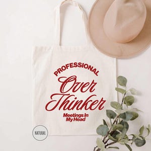 Pu&ograve; includere: Borsa tote color crema con la scritta "PROFESSIONAL Over Thinker Meetings In My Head" in rosso. Un cappello beige e un rametto di verde sono anche nell'immagine. La borsa ha un design semplice ed &egrave; realizzata in tessuto naturale.