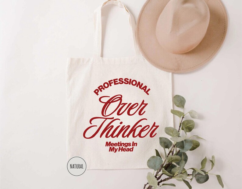 Pu&ograve; includere: Borsa tote color crema con la scritta "PROFESSIONAL Over Thinker Meetings In My Head" in rosso. Un cappello beige e un rametto di verde sono anche nell'immagine. La borsa ha un design semplice ed &egrave; realizzata in tessuto naturale.