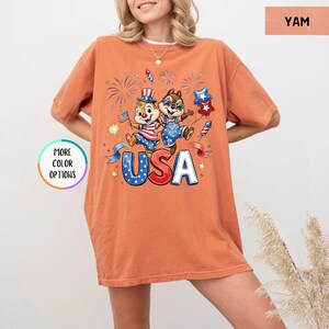 May include: Orange-colored t-shirt featuring a graphic of two cartoon chipmunks celebrating with fireworks, flags, and the word "USA" in blue and white. The shirt has short sleeves and a relaxed fit.