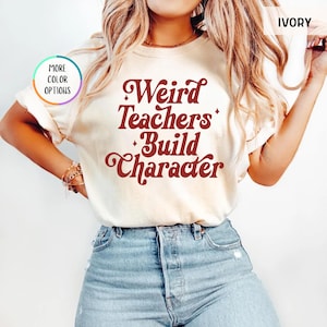 May include: Ivory-colored t-shirt with the text "Weird Teachers Build Character" in a retro font. The shirt is paired with blue jeans. The image also shows a "More Color Options" button.