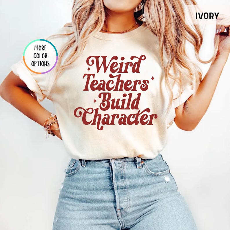 May include: Ivory-colored t-shirt with the text "Weird Teachers Build Character" in a retro font. The shirt is paired with blue jeans. The image also shows a "More Color Options" button.