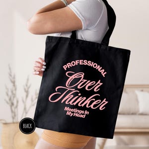 Pu&ograve; includere: Borsa tote nera con la scritta "PROFESSIONAL Over Thinker Meetings In My Head" in rosa. La borsa &egrave; tenuta da una persona che indossa una maglietta bianca e pantaloncini gialli. Un cerchio nero con la parola "BLACK" &egrave; nell'angolo in basso a sinistra.