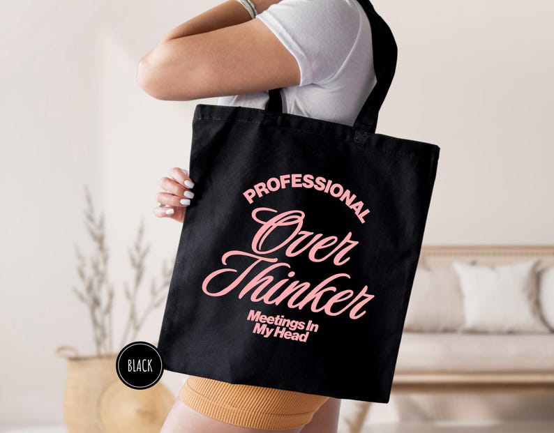 Pu&ograve; includere: Borsa tote nera con la scritta "PROFESSIONAL Over Thinker Meetings In My Head" in rosa. La borsa &egrave; tenuta da una persona che indossa una maglietta bianca e pantaloncini gialli. Un cerchio nero con la parola "BLACK" &egrave; nell'angolo in basso a sinistra.