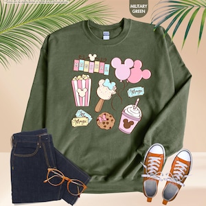 Puede incluir: Una sudadera verde con un dise&ntilde;o tem&aacute;tico de Mickey Mouse. El dise&ntilde;o incluye una pancarta que dice "MAGICAL", palomitas de ma&iacute;z, un helado de Mickey Mouse, una galleta de Mickey Mouse, un globo de Mickey Mouse y un batido de Mickey Mouse.